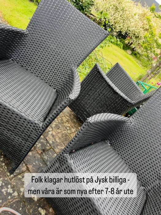 Svarta rottingstolar på en uteplats, nymålade och i bra skick efter flera år utomhus, med grön trädgård i bakgrunden.