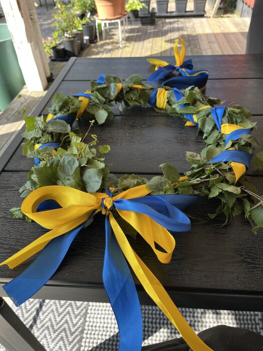 Midsommarkrans med björkgrenar och blå-gula sidenband ligger på ett svart bord utomhus.