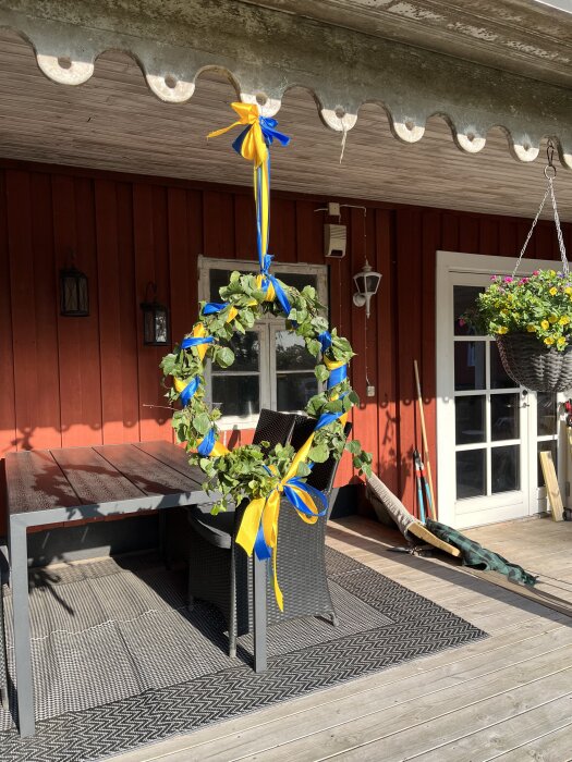 Midsommarkrans av björk och sidenband i blått och gult hänger på altan med rödmålad vägg samt utemöbler under tak.