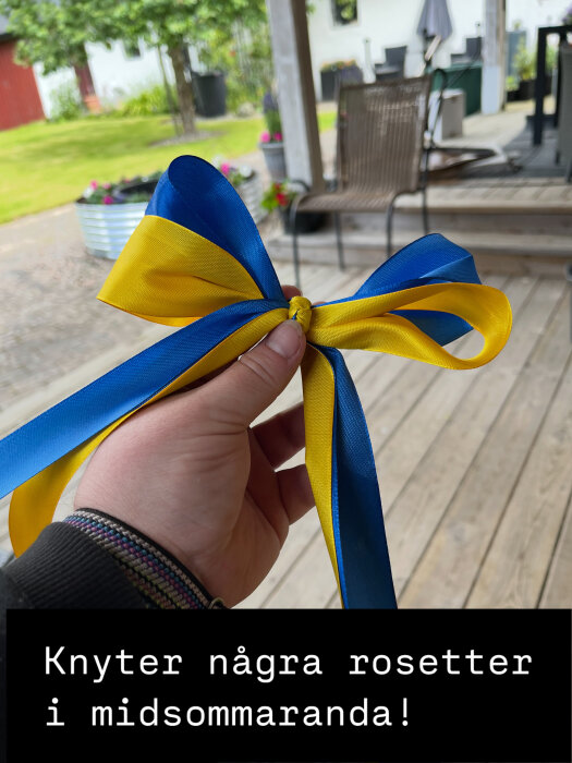 En hand håller en blågul rosett i en trädgård med en uteplats i bakgrunden.