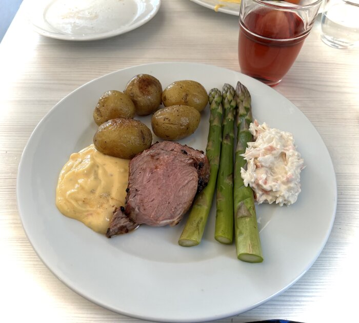 Tallrik med grillat kött, kokt sparris, ugnsstekta potatisar, coleslaw och bearnaisesås, med ett glas röd dryck bredvid.
