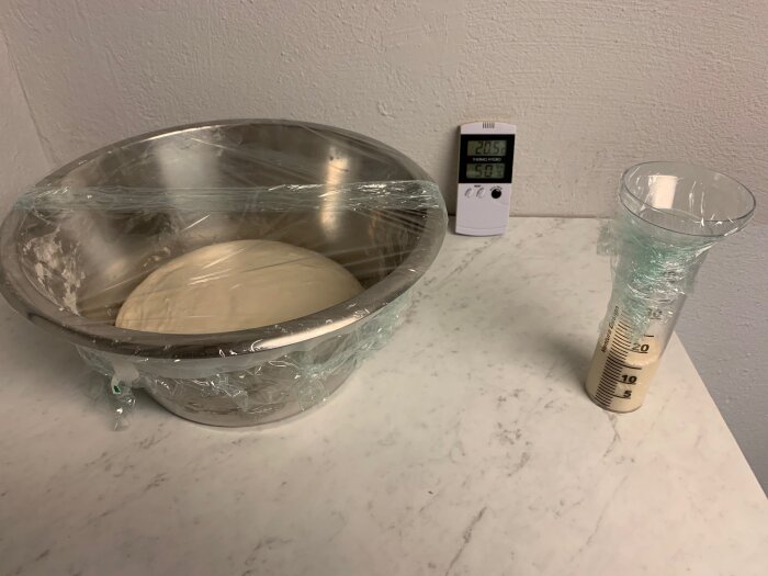 Deg på jäsning i en täckt metallbunke bredvid en graderad cylinder på ett marmorbord. I bakgrunden en digital termometer.