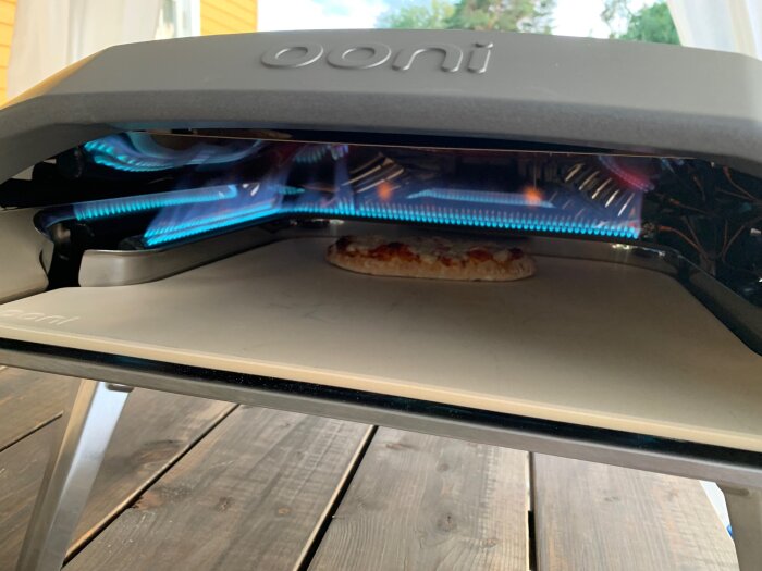 Utomhus gasugn med brinnande lågor, en pizza inuti på baksten, stående på ett träbord.