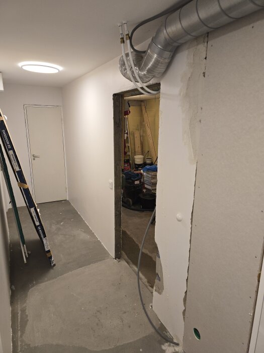 Korridor i källare med nylagd betonggolv, en rivet väggparti och synliga ventilationsrör som går genom väggen, dörröppning till hobbyrum.