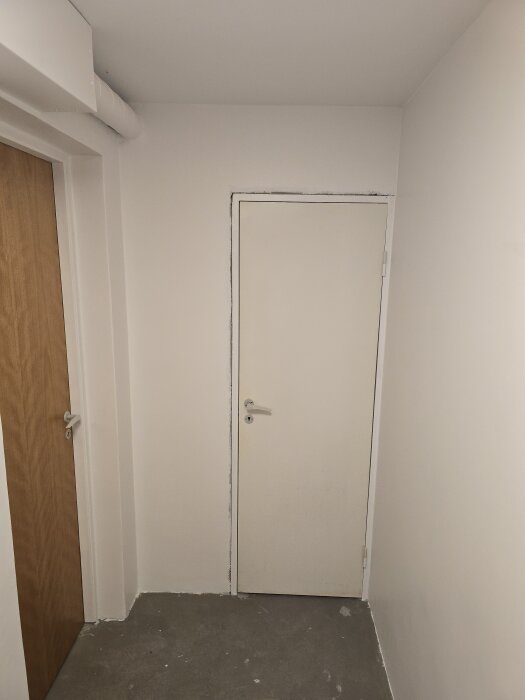 Vit dörr i ett nygipsat rum med en gråmålad betonggolv. Bredvid syns en brun dörr och ett ventilationsrör överst i vänstra hörnet.