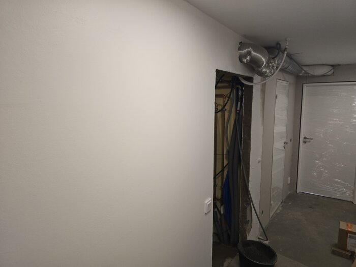 Väggen i en källare under rivning med synliga rör och ventilationssystem i taket, samt en dörr täckt med plast i bakgrunden.