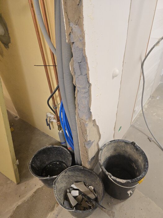 Tre svarta hinkar fyllda med byggrester vid en delvis riven innervägg av murad blåbetong i en källare under renovering.