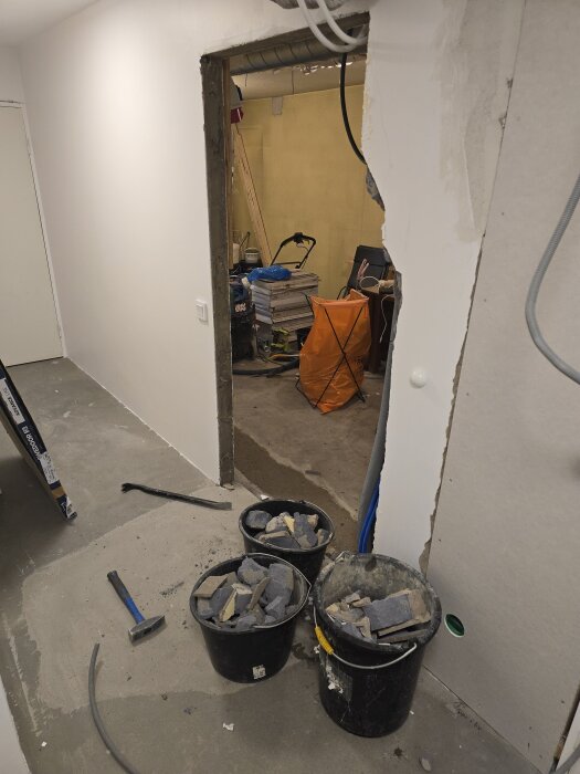 Tre hinkar fyllda med rivna betongbitar vid en delvis riven vägg, i en källarkorridor under renovering. Ett verktyg ligger bredvid.