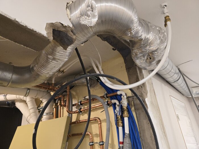 Rör och kablar i en halvfärdig vägg med flexibla ventilationsledningar och diverse kopplingar i en källare under renovering.