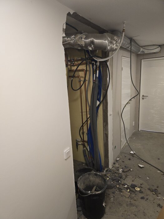 Rivningsprojekt i källare med trasig vitputsad vägg, synliga ventilationsrör och sladdar, svart hink för byggskräp på golvet.