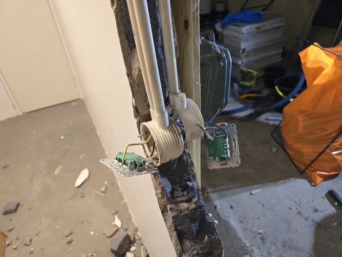 Närbild på en riven vägg i blåbetong med två eldosor ihopspikade rygg mot rygg, kablar och rör synliga mellan murverket. Verktyg och material i bakgrunden.