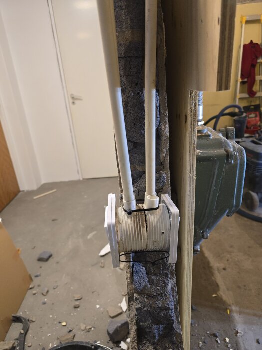 Riven murad blåbetongvägg med synliga VP-rör och elkopplingar, belyst av arbetslampa i källaren under renovering.