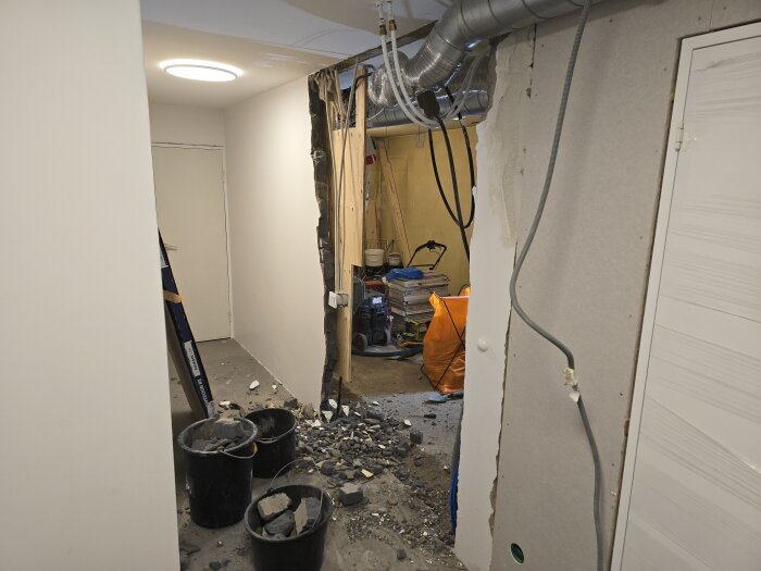 Rivningsarbete på icke-bärande vägg av blåbetong i källare, med tre bygghinkar fyllda med rivningsmaterial och synliga VP-rör och ventilationsrör.