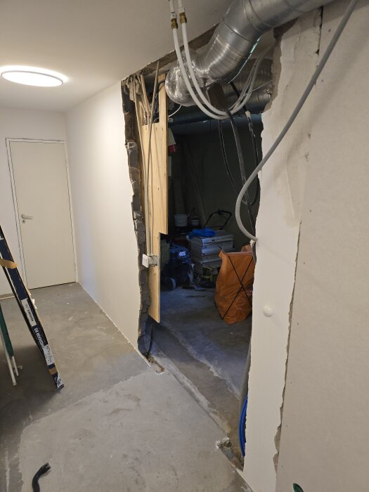 Riven vägg i källare, blottar rör och elkablar; synliga ventilationsrör och nygjuten betong på golv. Doorway till hobbyrum under renovering.