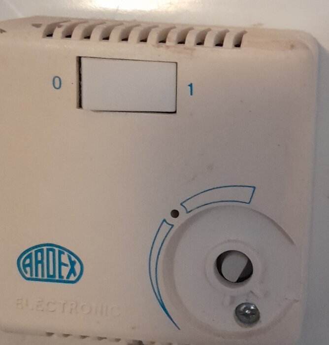 Termostat med vit panel och ratt, märkt "ARDEX," visar inställningar 0 och 1. Ratthålet är synligt med skruv under, vilket avslöjar insidan.