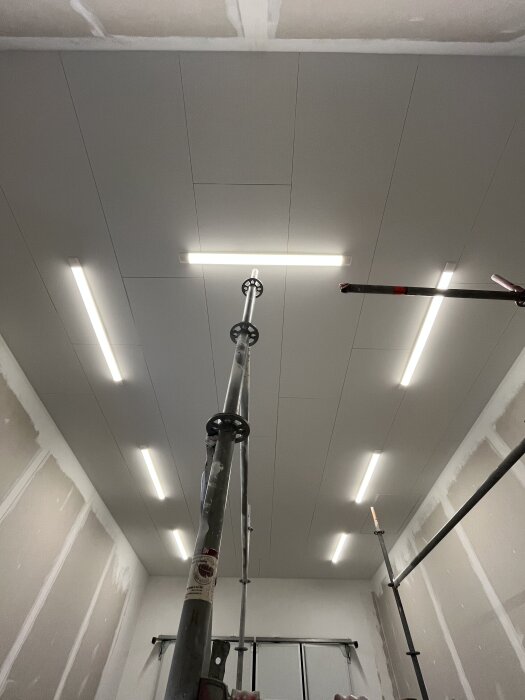 Takbelysning i ett nyrenoverat rum med plankor i taket och väggarnas grundarbete synligt. Byggnadsställning i mitten av rummet.