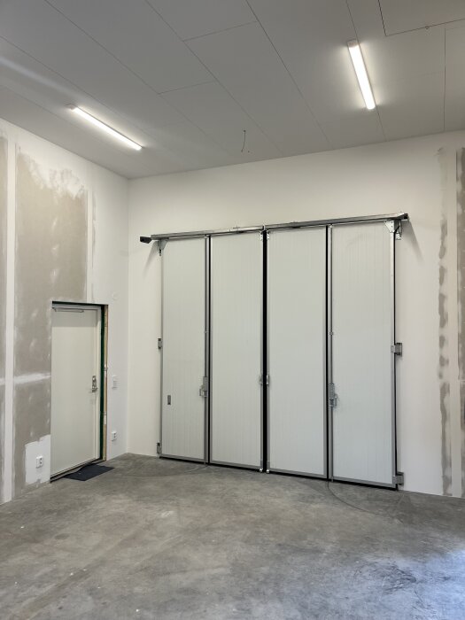 Nyligen elektrifierad och tom byggnadsinteriör med vita väggar, betonggolv, stora vikdörrar och lysrör i taket.