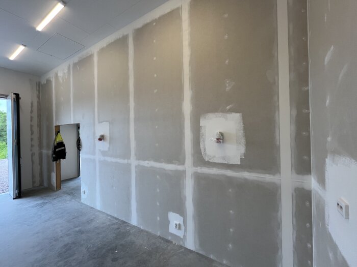 Nybyggt rum med spacklade väggar, installerade eluttag och lysande taklampor. Dörr till vänster visar ett utomhusområde.