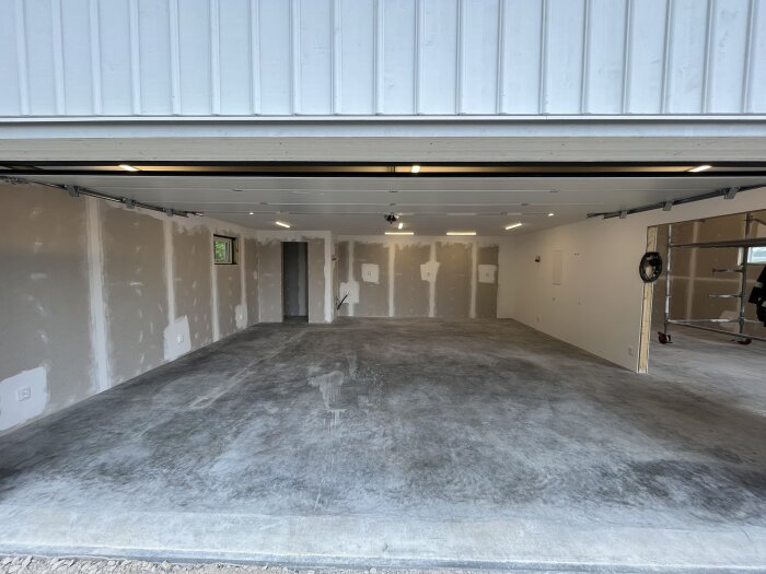 Tomt garage med vita väggar och betonggolv, taklampor och en öppen garageport. Viss byggställning och kablar synliga.