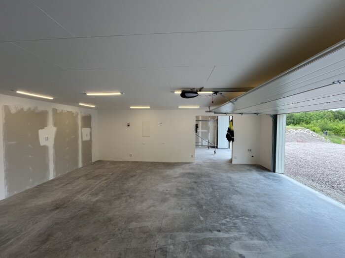 Tomt garage med betonggolv, vitmålade väggar och öppnad garageport. Flera lysrör i taket. Sedd från insidan ut mot grönska.