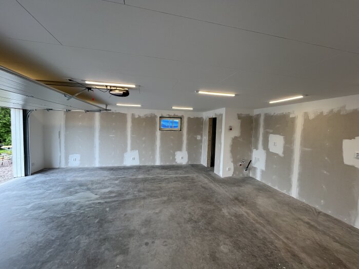 Tomt garage med vitmålade betongväggar, taklampor och oavslutade väggar. Fönster och garageport syns, inga möbler eller inredning.