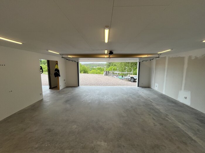 Nybyggt garage med betonggolv och öppna portar som visar grusuppfart utanför, minimalistisk inredning och takbelysning.