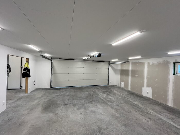 Tomt garage med betonggolv, elbelysning och en garageport, delvis ofärdig vägg med gipsplattor.