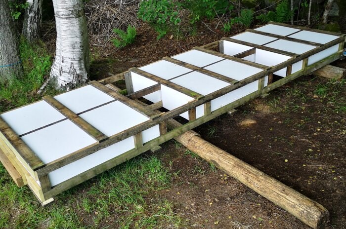 En flytbrygga under renovering med vit flytmaterial infällt i en träram, bredvid träd och markvegetation.