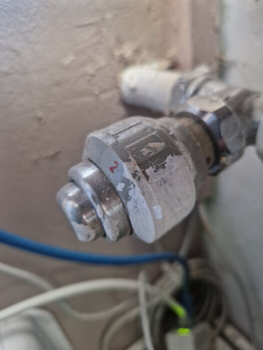 Gammal vitmålad termostatventil med metallmutter och plastdetaljer installerad i ett hus, med vägg och kablar i bakgrunden.