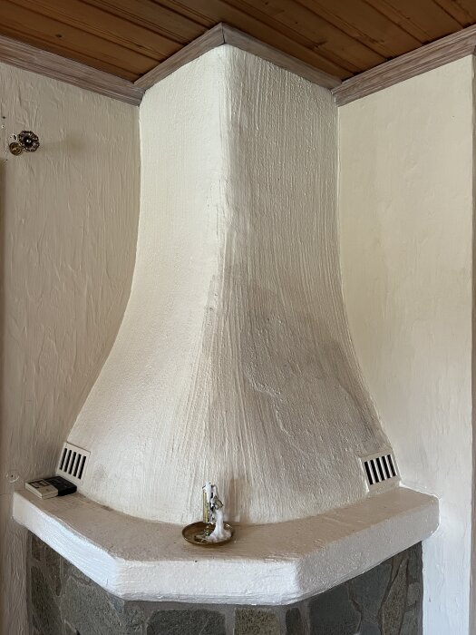 Vitmålad murstock med grov struktur och ventilationsgaller samt en ljushållare på murstockens kant. Tak i träpanel ovanför.