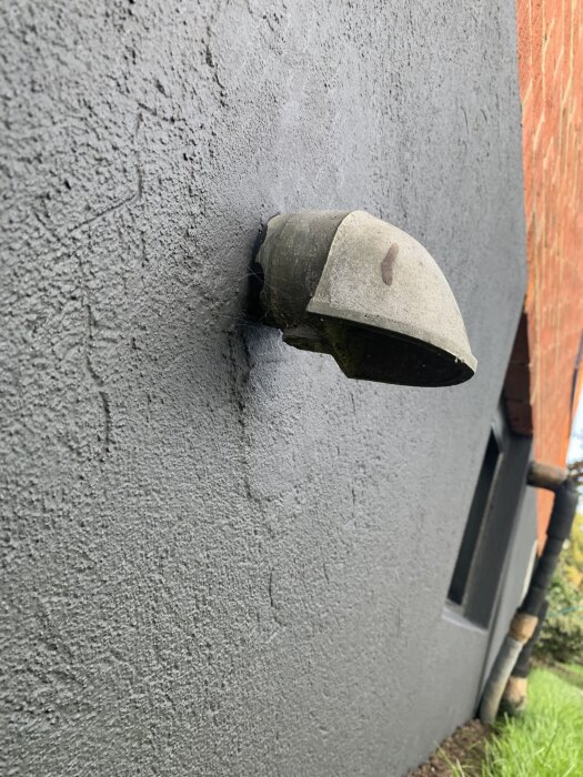 Ventilhuv på grå putsad vägg, fäst på ett utstickande rör, nära en tegelvägg och grön gräsmatta.