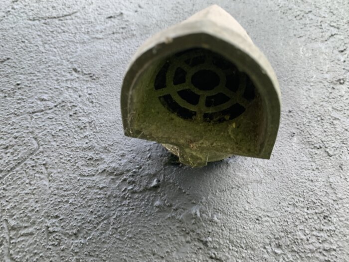 Metallisk del med ventilerad öppning, monterad på en yta, används på ett 60 mm rör genom vägg. Okänd beteckning.