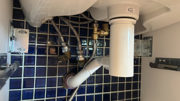 Vattenlås och rörinstallation under handfat mot blå kakelvägg, inkluderar blandarventiler och flexibla anslutningsrör.