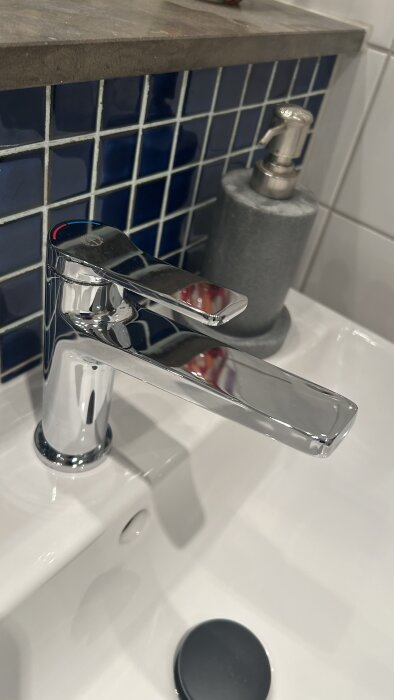 Kromad tvättställsblandare av märket Gustavsberg installerad på vitt handfat med blå kaklade väggar i bakgrunden.