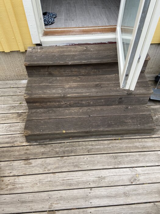 Mörk trapp i trä med bredvidliggande ljusare altan. Dörren är öppen, vilket visar ett ljust golv inomhus.