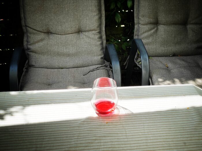 Glas med röd dryck på ett randigt bord framför två gråa utomhusstolar med dynor.