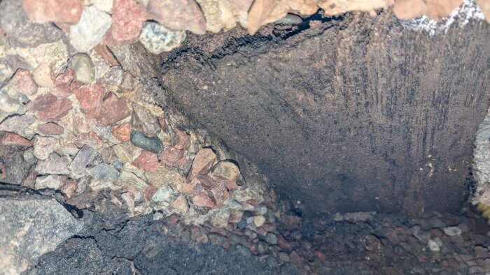 Hål under betongplatta med jord och grus synliga, delvis belyst med LED-lampa, potentiella ruttnande trädelar uppmärksammas.