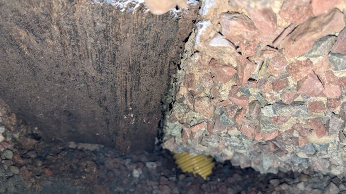 Bild av ett hål under en platta med synliga stenar och jord; potentiellt ruttnande trä samt ljus belysning i nedre delen.