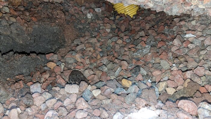 Ett hål under en husgrund med synliga stenar och grus, markerad av LED-ljus som skapar svarta linjer. Syns även ett gult rör.