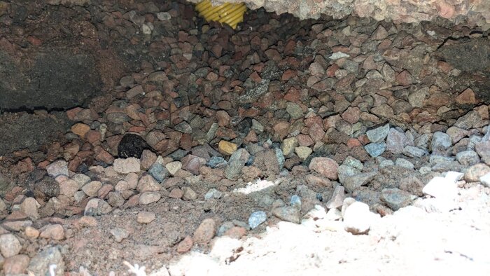 Stort hål under husgrund visar stenar och grus, delvis upplyst. En gul slang syns längst in under plattan.