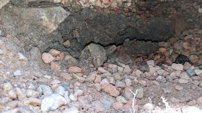 Hål under huset med små stenar och jord synliga. Hålet sträcker sig in under plattan och är omringat av stenigt och mörkt material.