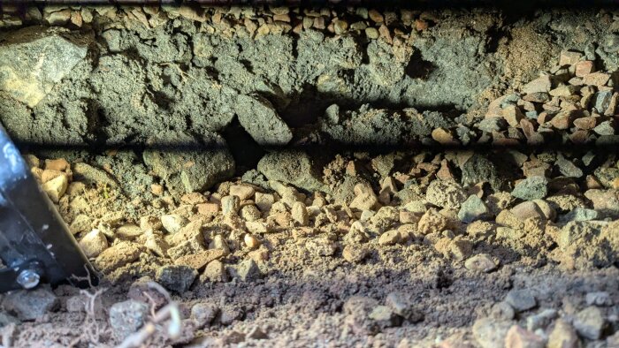 Hål i marken invid platta med synliga stenar och jord, belyst delvis med LED-lampa; svarta skugglinjer syns över bilden.