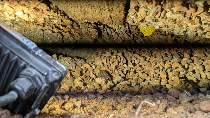 Grus och stenar under en husgrund med belyst hål, svart lampa delvis synlig till vänster.