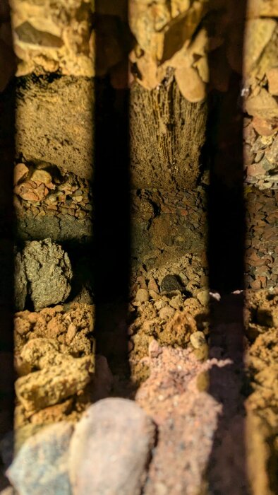 Mörkt hål under husgrund med jord och grus, belyst av LED-lampa, samt svarta skuggor från lampan synliga på bilden.