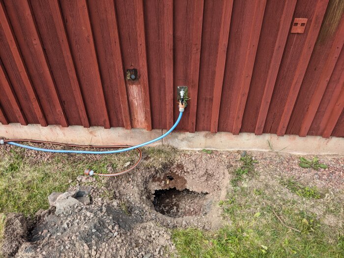 Hål i marken framför en röd träfasad med vattenutkastare och blå slang. Gräset runt om är delvis avlägsnat och jorden är exponerad.