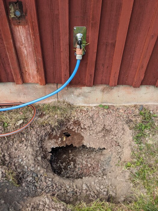 Hål i marken vid husvägg med vattenutkastare, visar jord och grus. Blå slang kopplad till vattenutkastaren. Gräs och jord omger hålet.
