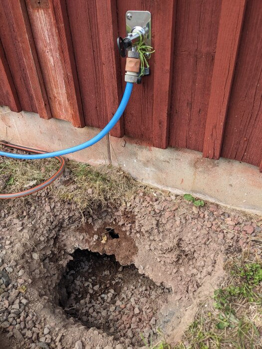 Hål i marken under en blå slang kopplad till en vattenutkastare på en träfasad. Jorden runt hålet är uppluckrad och det finns grus inuti hålet.