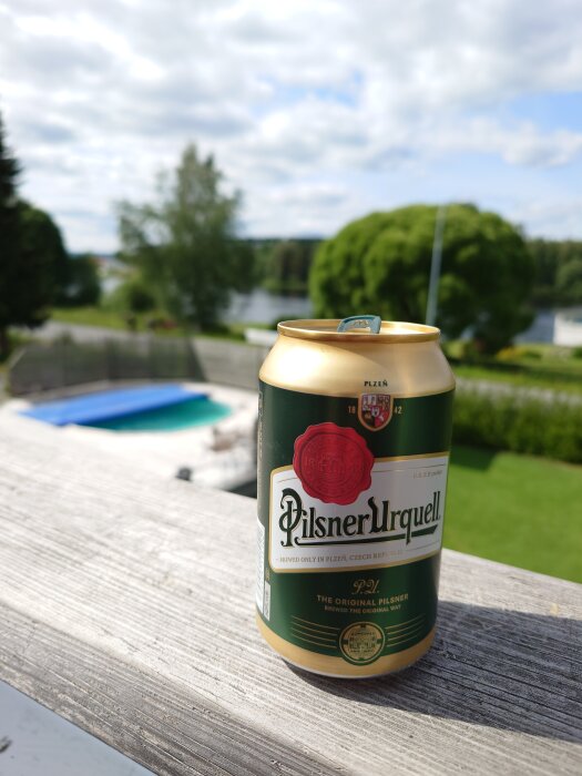 En burk Pilsner Urquell står på ett altanräcke med en utsikt över en trädgård, pool och en sjö i bakgrunden under en molnig himmel.