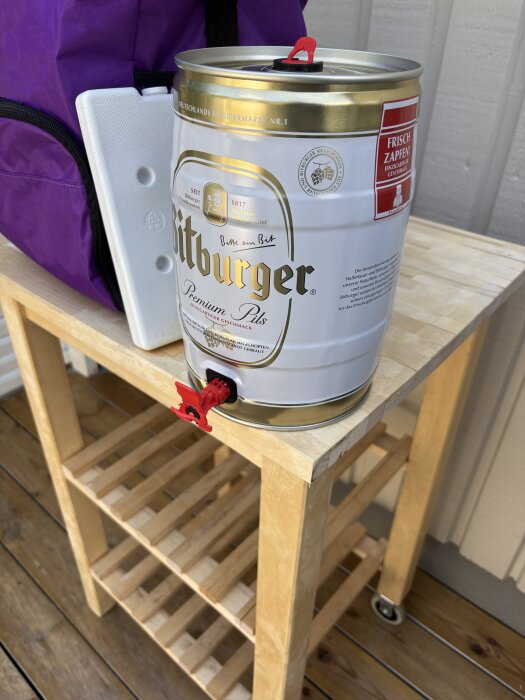 Mini-fat med öl placerat på en trävagn, bredvid en lila väska och en vit kylklamp.