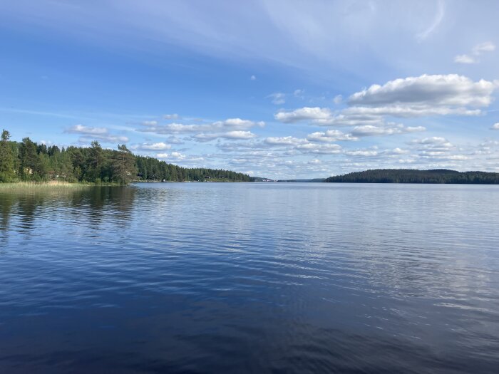 Sjölandskap med klarblå himmel, vita moln och skogsklädda stränder.